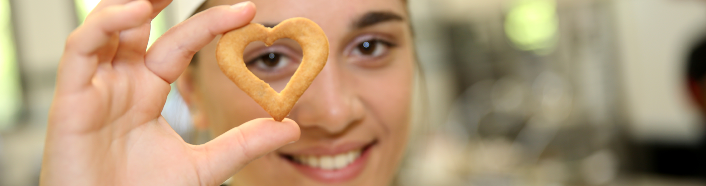Eine junge Frau hält lächelnd einen Keks in Herzform vor ihr rechtes Auge.